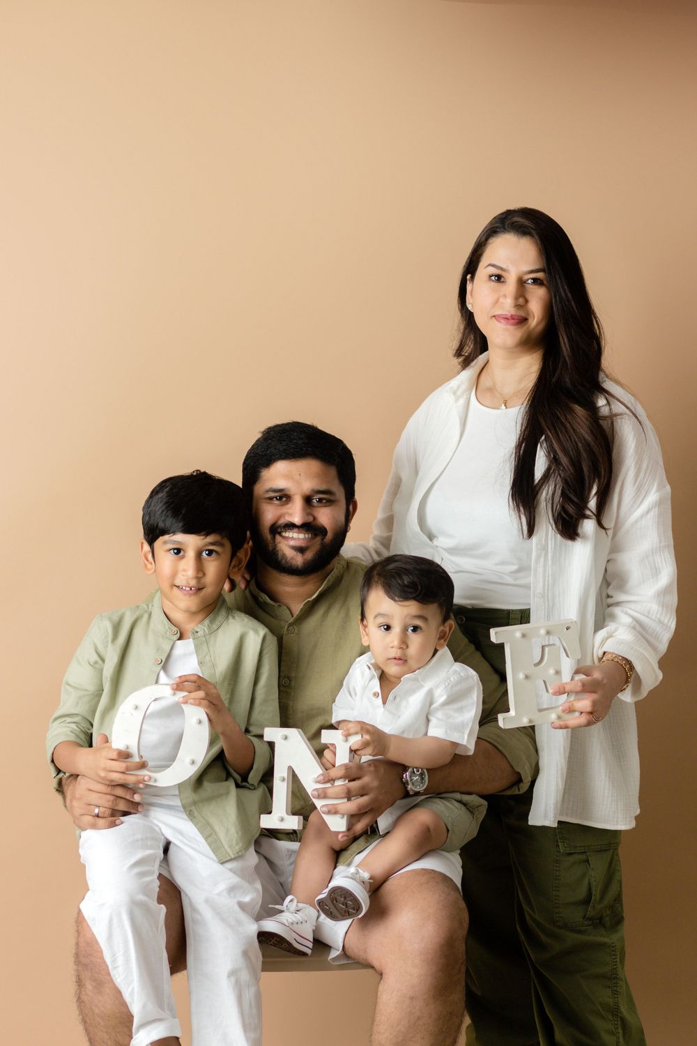 Family of four in coordinated white and olive green outfits posing against beige background.