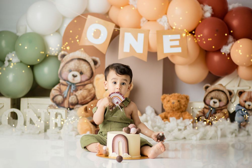 Baby's first birthday setup with teddy bears and peach balloon arch displaying 'ONE' banner.