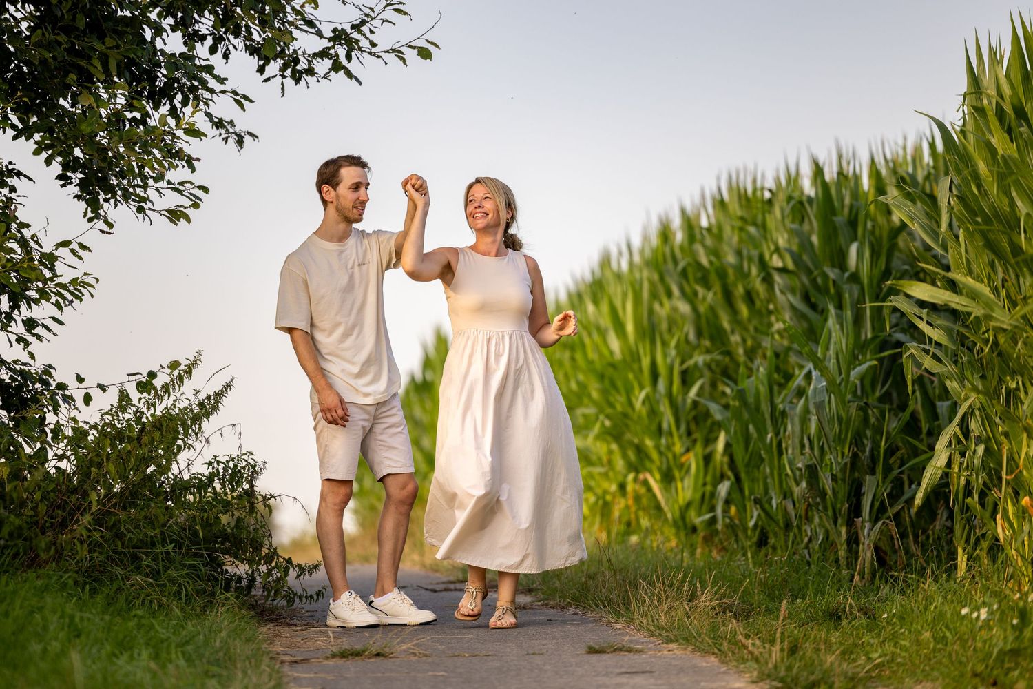 A couple in light summer clothing walking together on a path beside a cornfield at sunset.