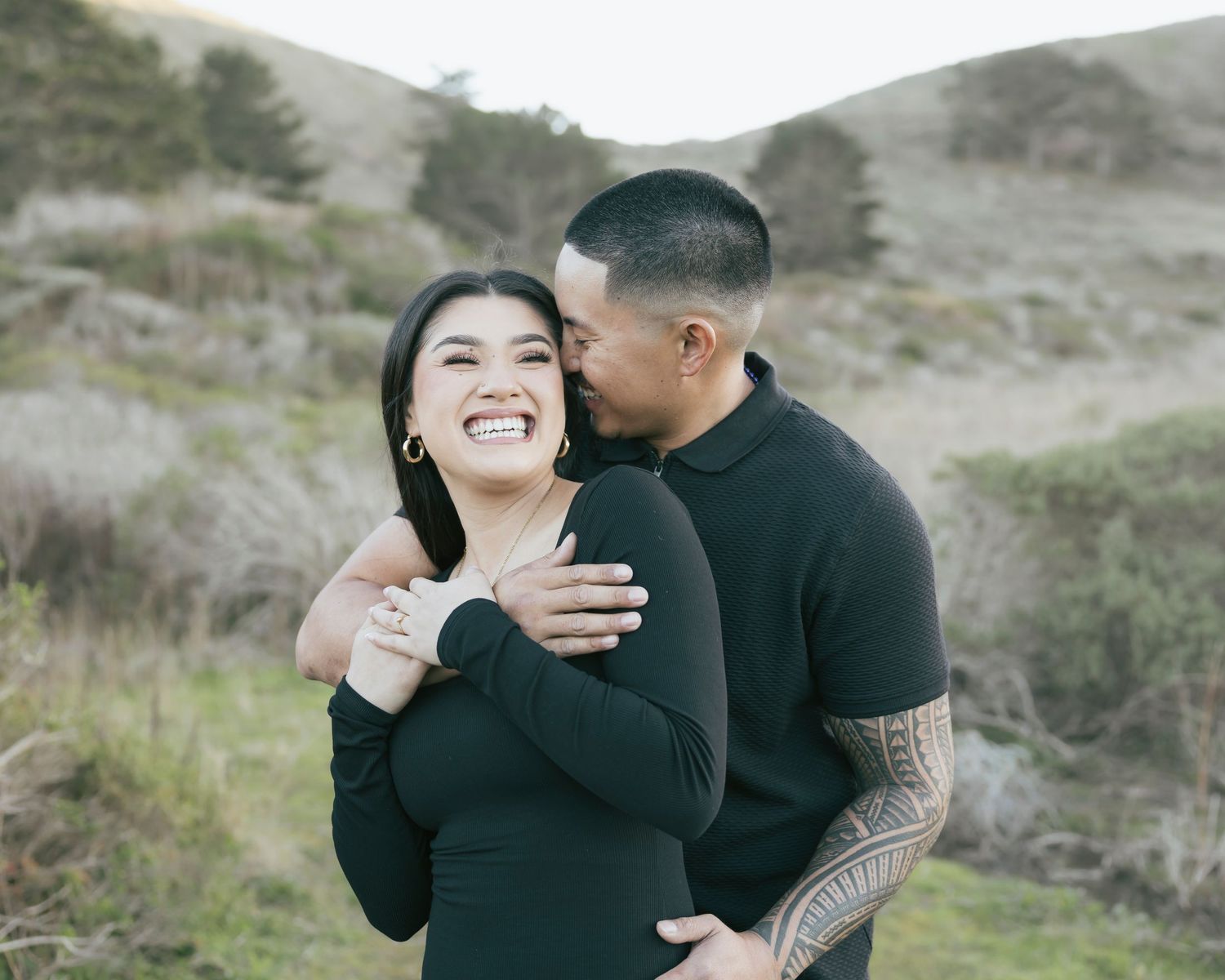 A couple in black sweaters share an intimate moment outdoors against a backdrop of rolling hills and brush.