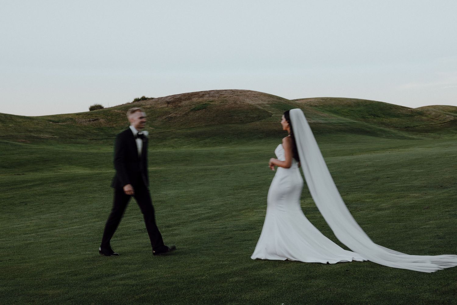 A bride with flowing veil and groom walk across a rolling green golf course.