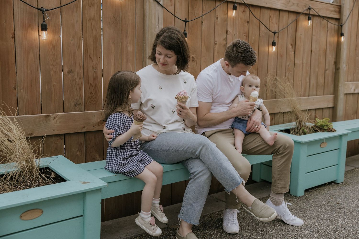 A family relaxes together on mint green wooden benches in a backyard with string lights and wooden fence backdrop.