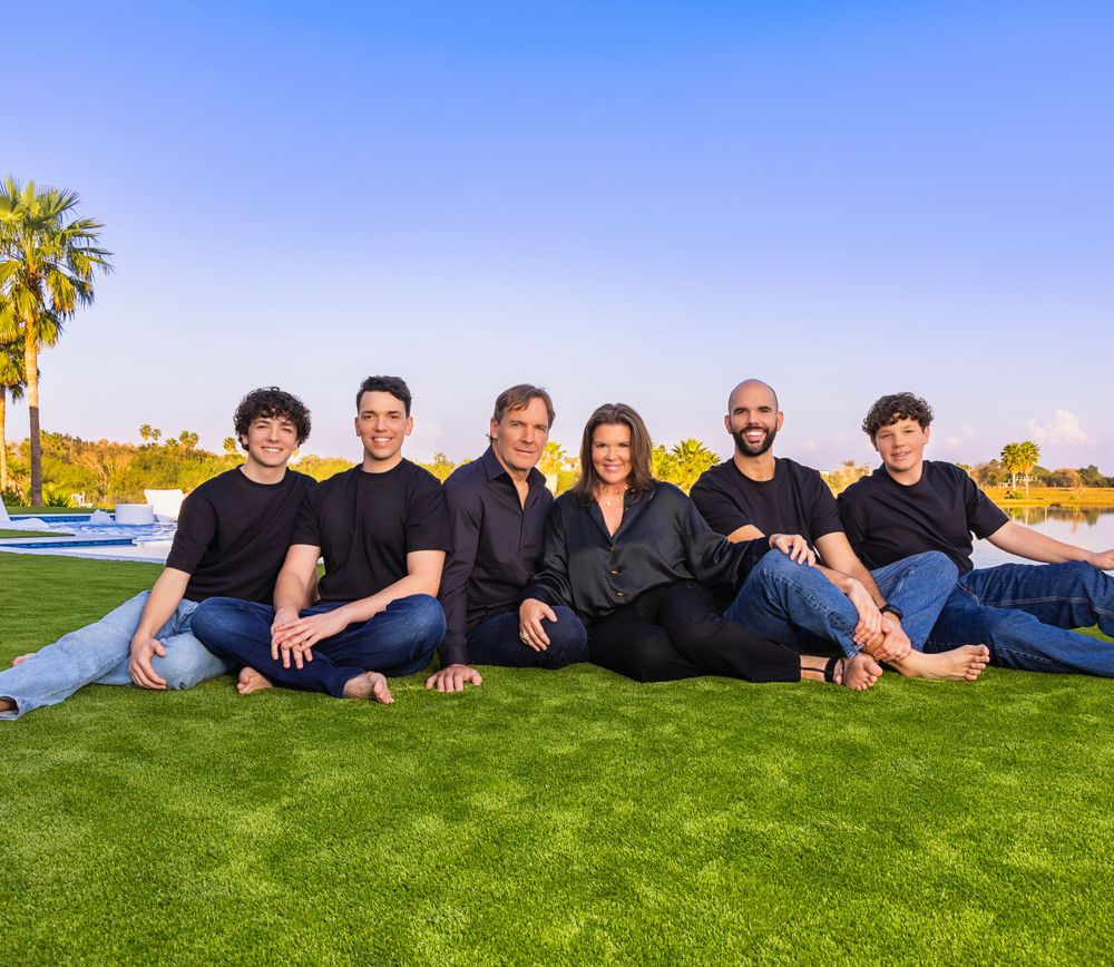 A group of people wearing black clothing sit together smiling on green grass with palm trees and blue sky in the background.