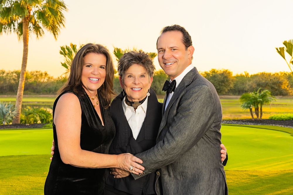 Three people in formal attire posing together on a golf course at sunset with palm trees in the background.
