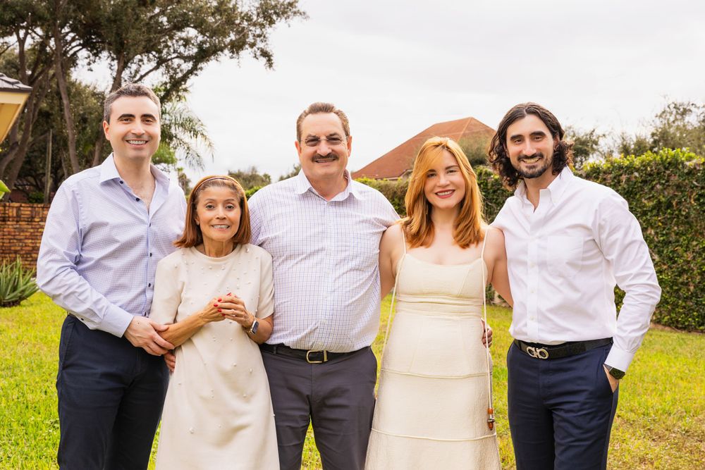 Group of people dressed in light summer attire posing together on a sunny lawn with trees and buildings in background.