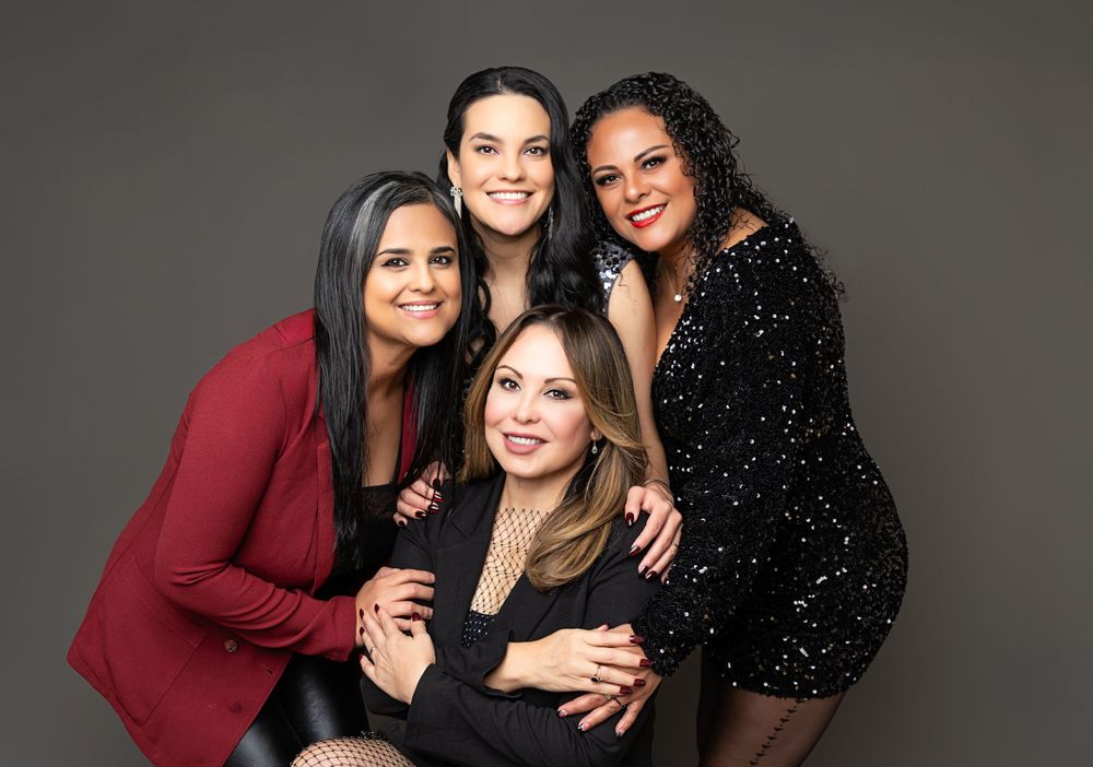 Four women pose together for a studio portrait, wearing a mix of red and black outfits against a gray background.