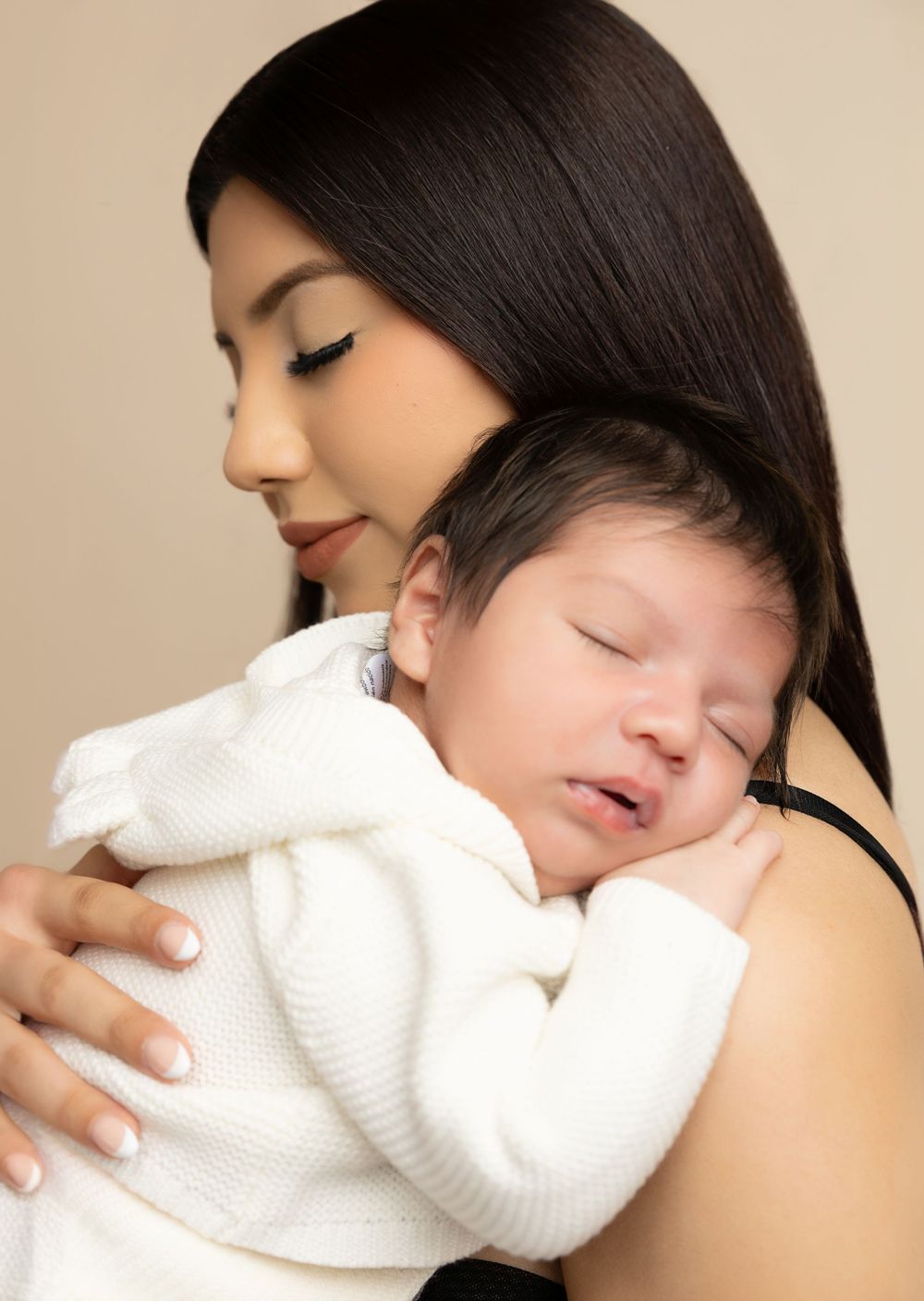 Tender moment between mother and sleeping newborn baby in white sweater.