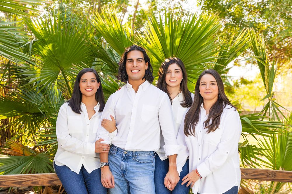 A group in matching white shirts poses together in front of tropical palm plants on a sunny day.