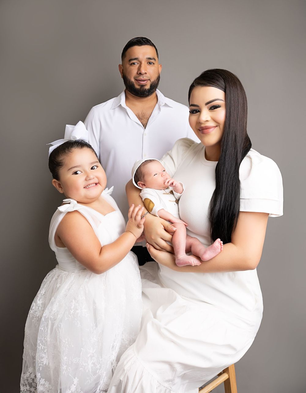 A family of four wearing white poses together for a studio portrait with their newborn baby.