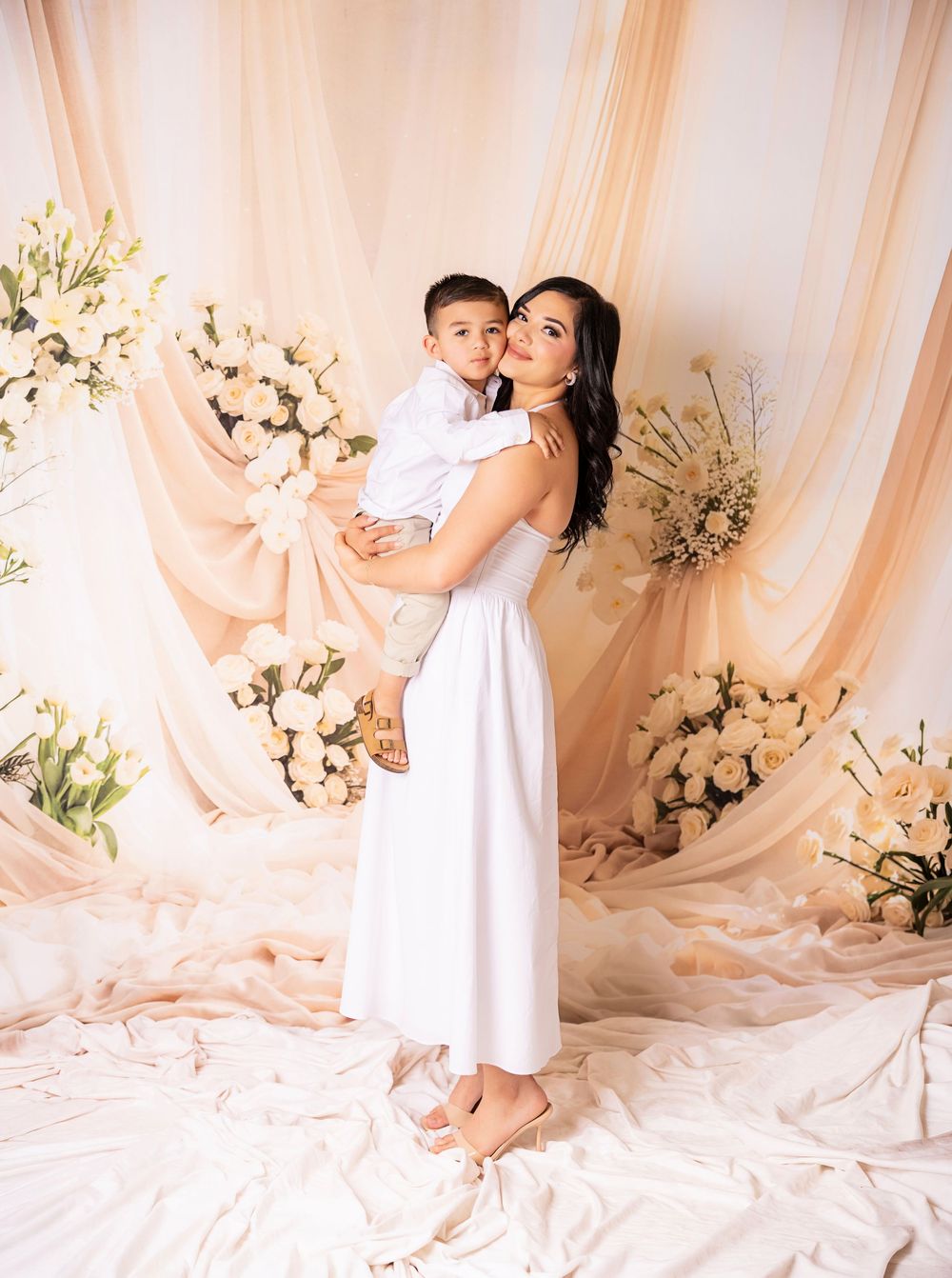 Woman in white dress holds a young boy, posing before a floral backdrop with cream draping and white roses.