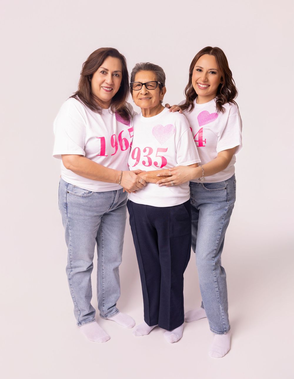 Three generations of women in matching pink and white shirts pose together for a breast cancer awareness photo.