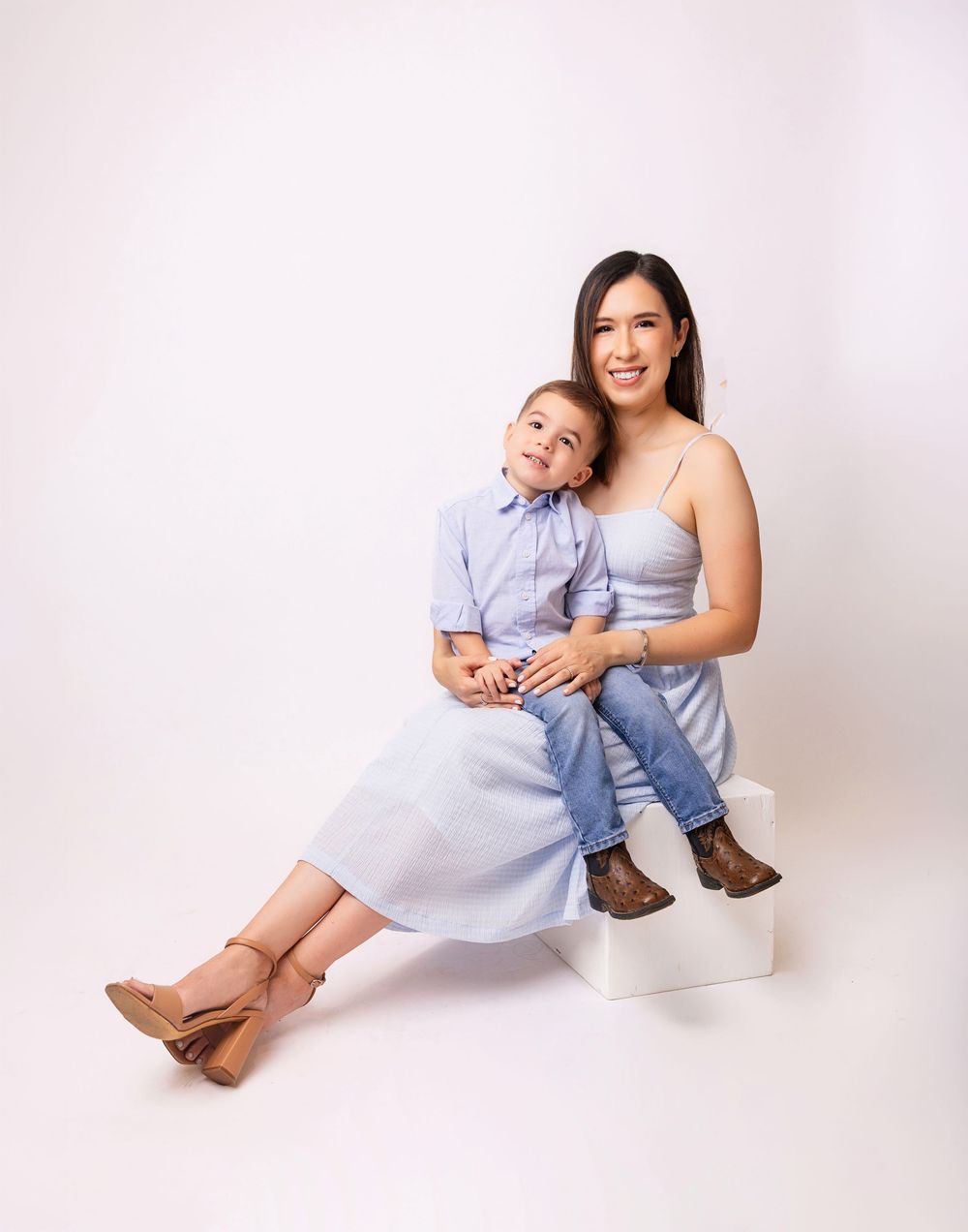 A smiling woman in a light blue dress sits with a young boy in a blue shirt and jeans on a white cube.
