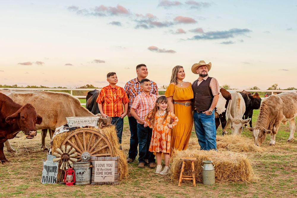 Group gathers at sunset for rustic western photoshoot with hay bales and vintage wagon wheel in golden field.