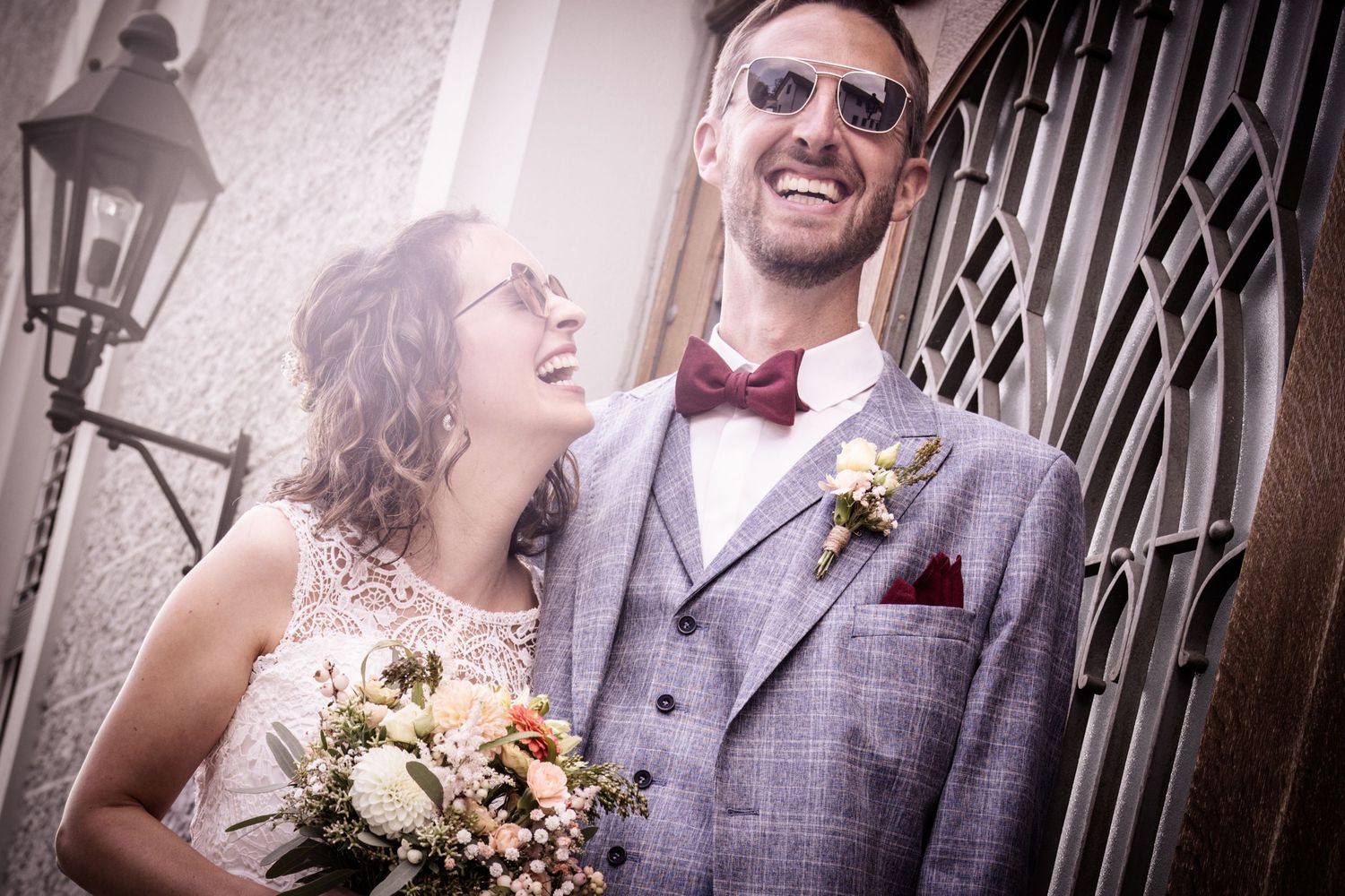 A joyful wedding couple laughing together outside an ornate doorway, with the bride holding a bouquet of flowers.