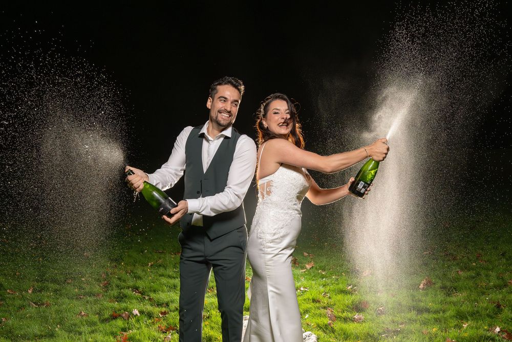 A couple in wedding attire spraying champagne bottles outside at night with backlit mist creating a magical effect.