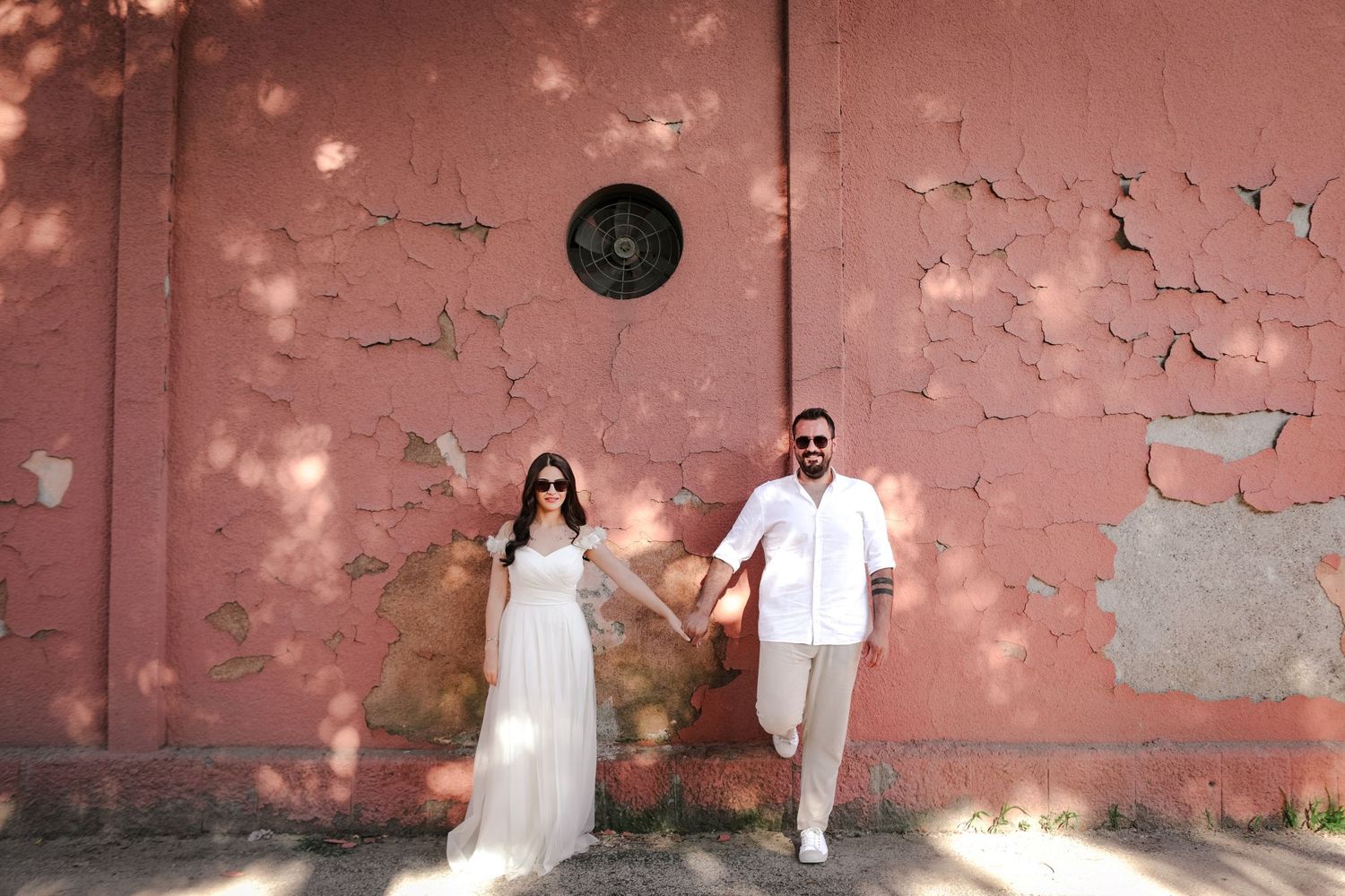 A couple stands against a weathered pink wall in white outfits for a stylish photo.