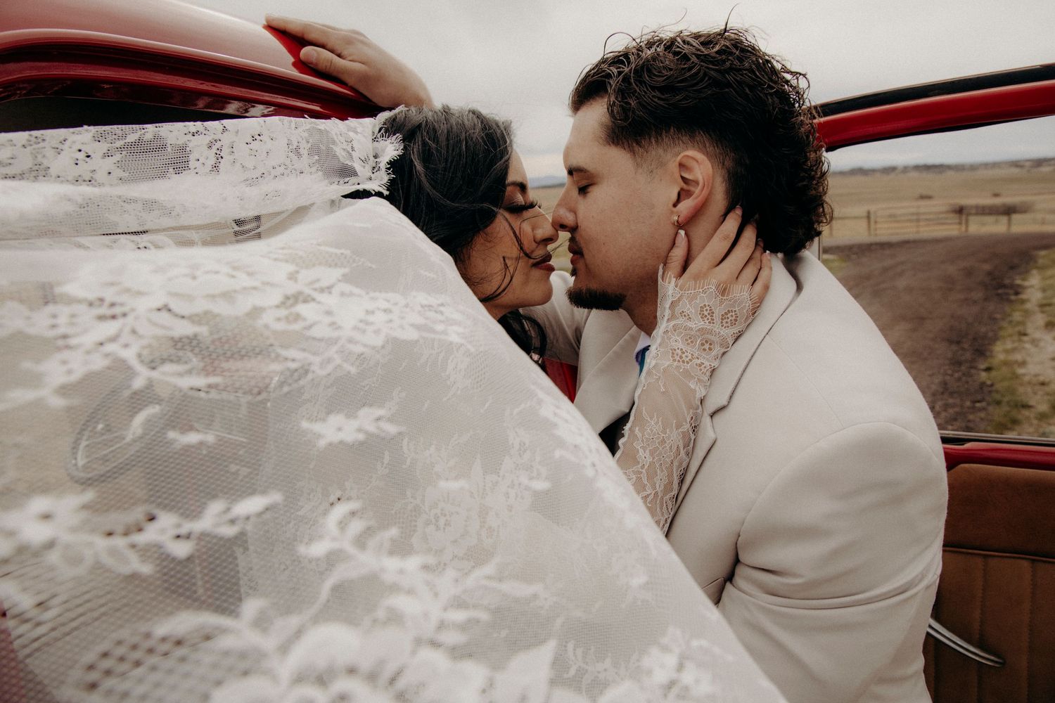 Bride and groom share a romantic kiss beside a red vintage vehicle on their wedding day.