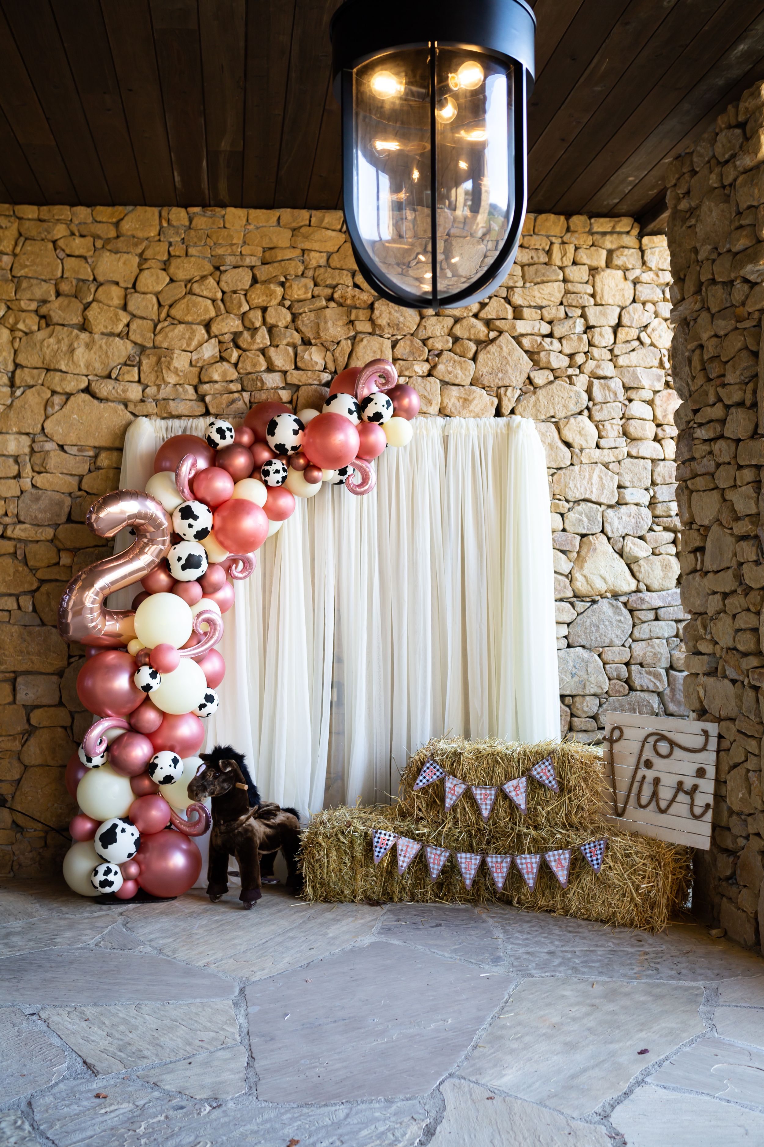 Party decoration setup with pink balloon arch, hay bales, and white curtain backdrop.