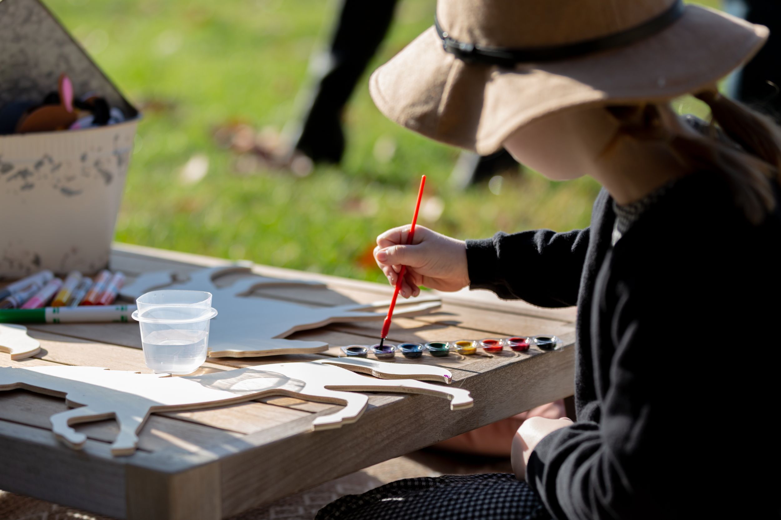 Person wearing dark clothing and hat paints with watercolors at outdoor table with art supplies.
