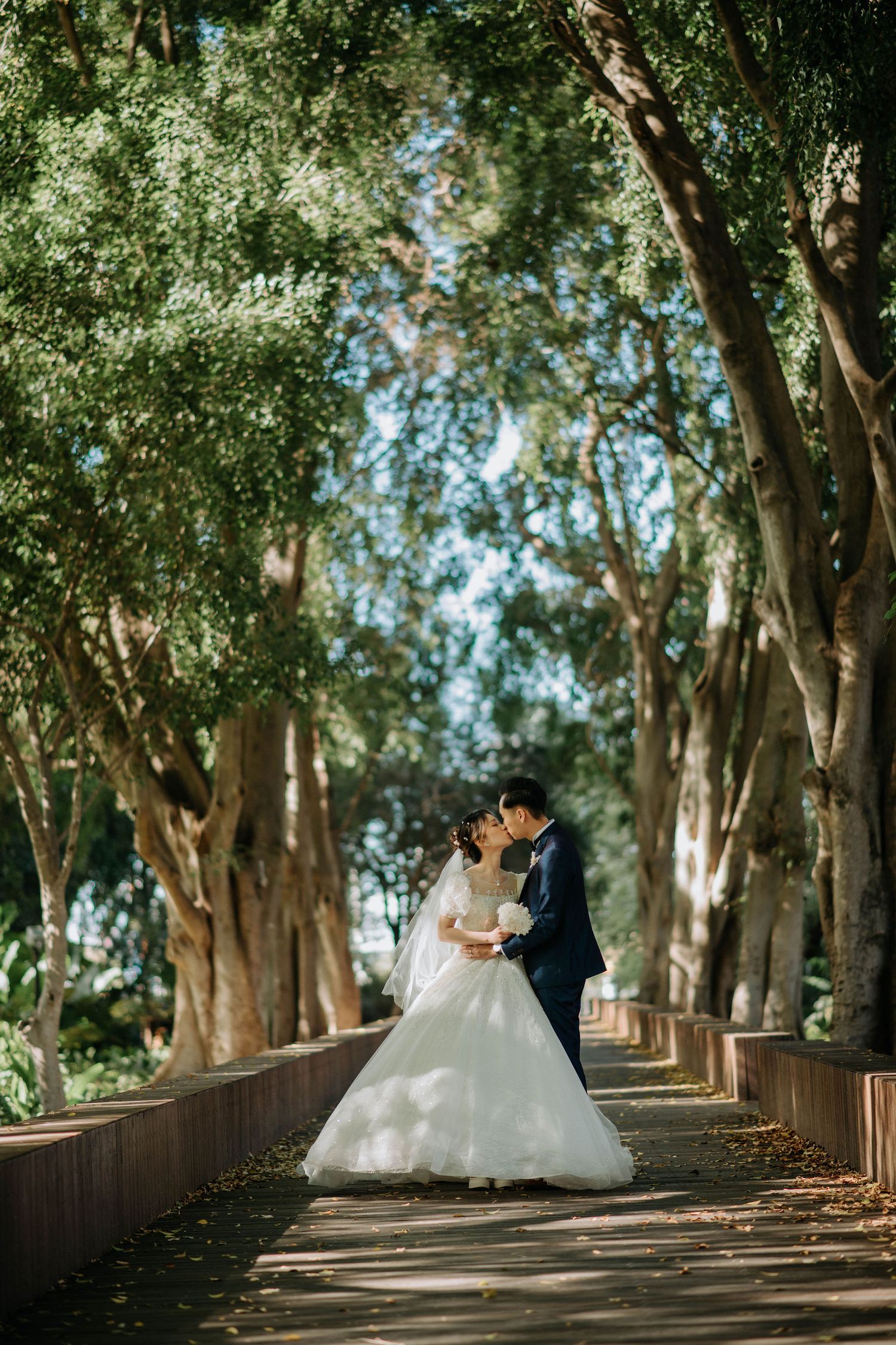 Couple in wedding attire share romantic moment on tree-lined pathway.
