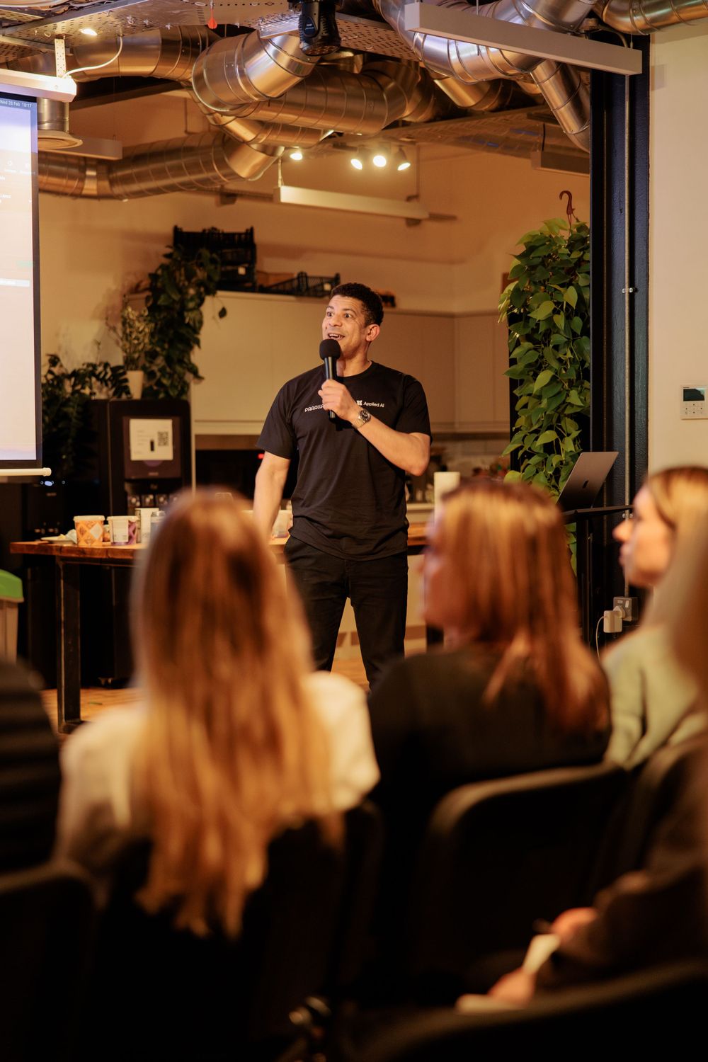 Speaker presenting to seated audience in modern venue with indoor plants.