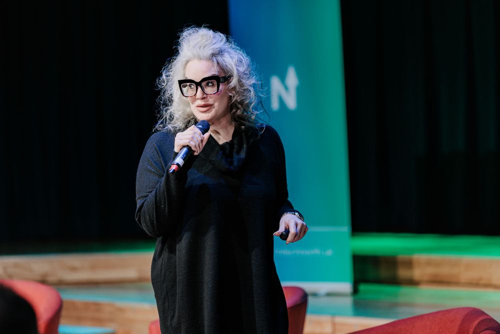 Speaker in black outfit presenting against green backdrop during conference.