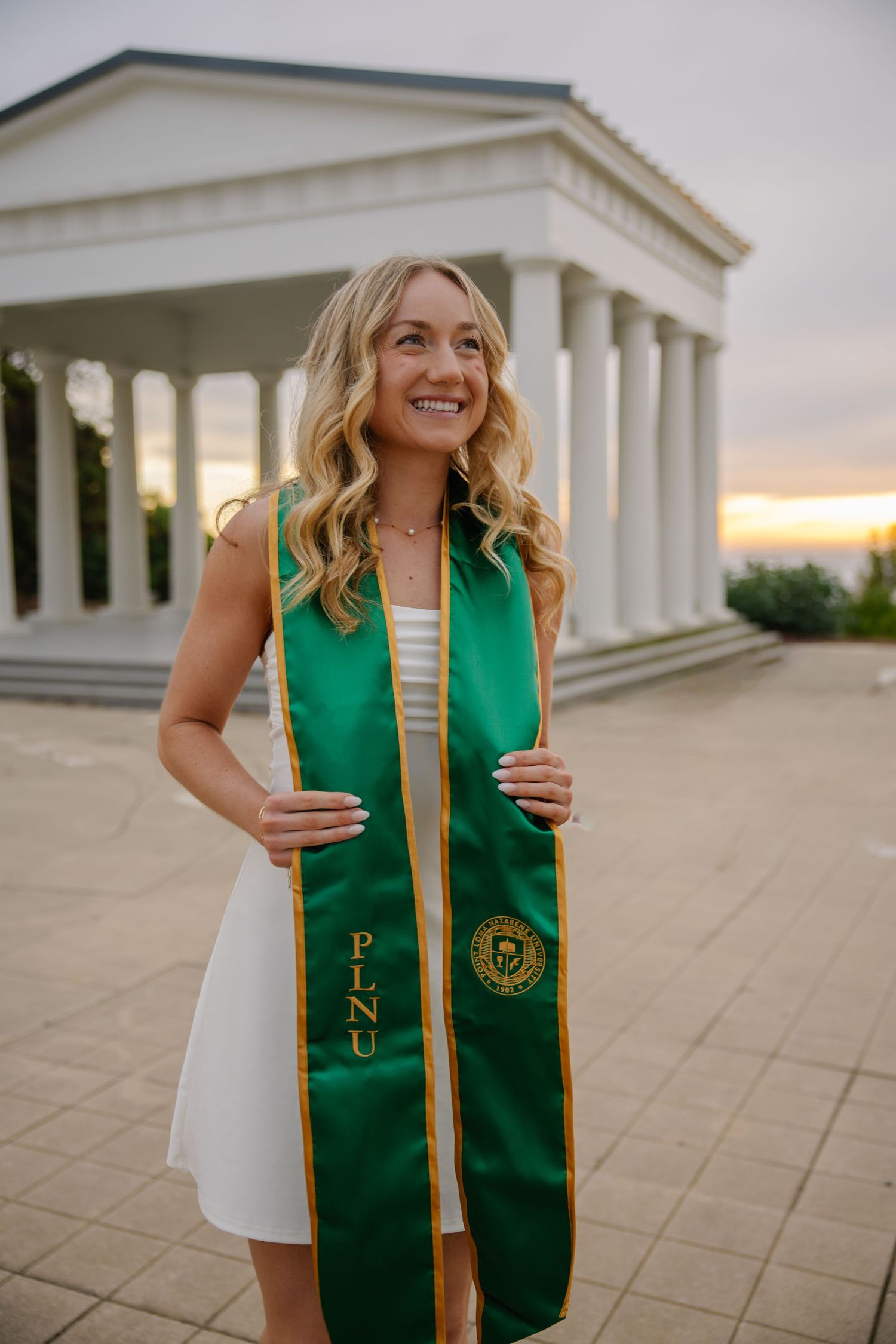 A graduate in white dress and green stole poses in front of a classical building at sunset.