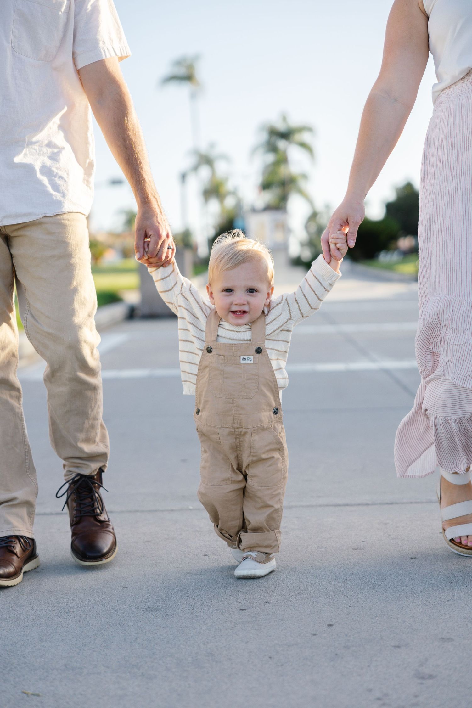 Toddler in overalls walking while holding parents' hands on sunny sidewalk with palm trees.