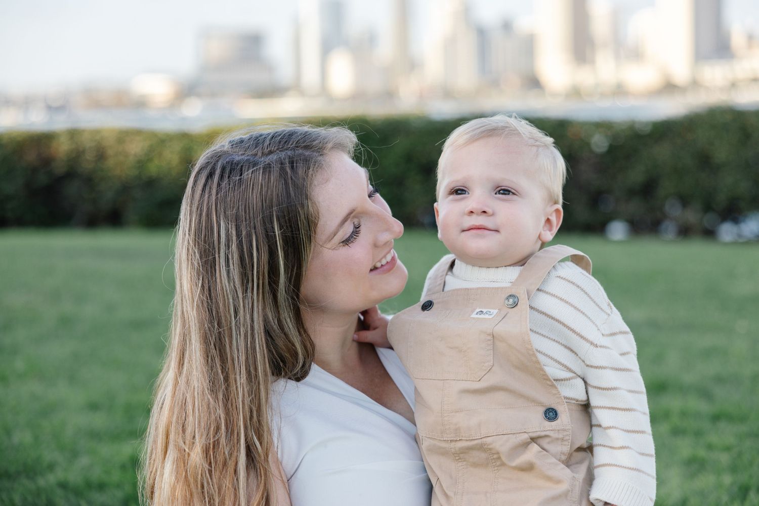 Mother and baby in overalls share sweet moment with cityscape in background.