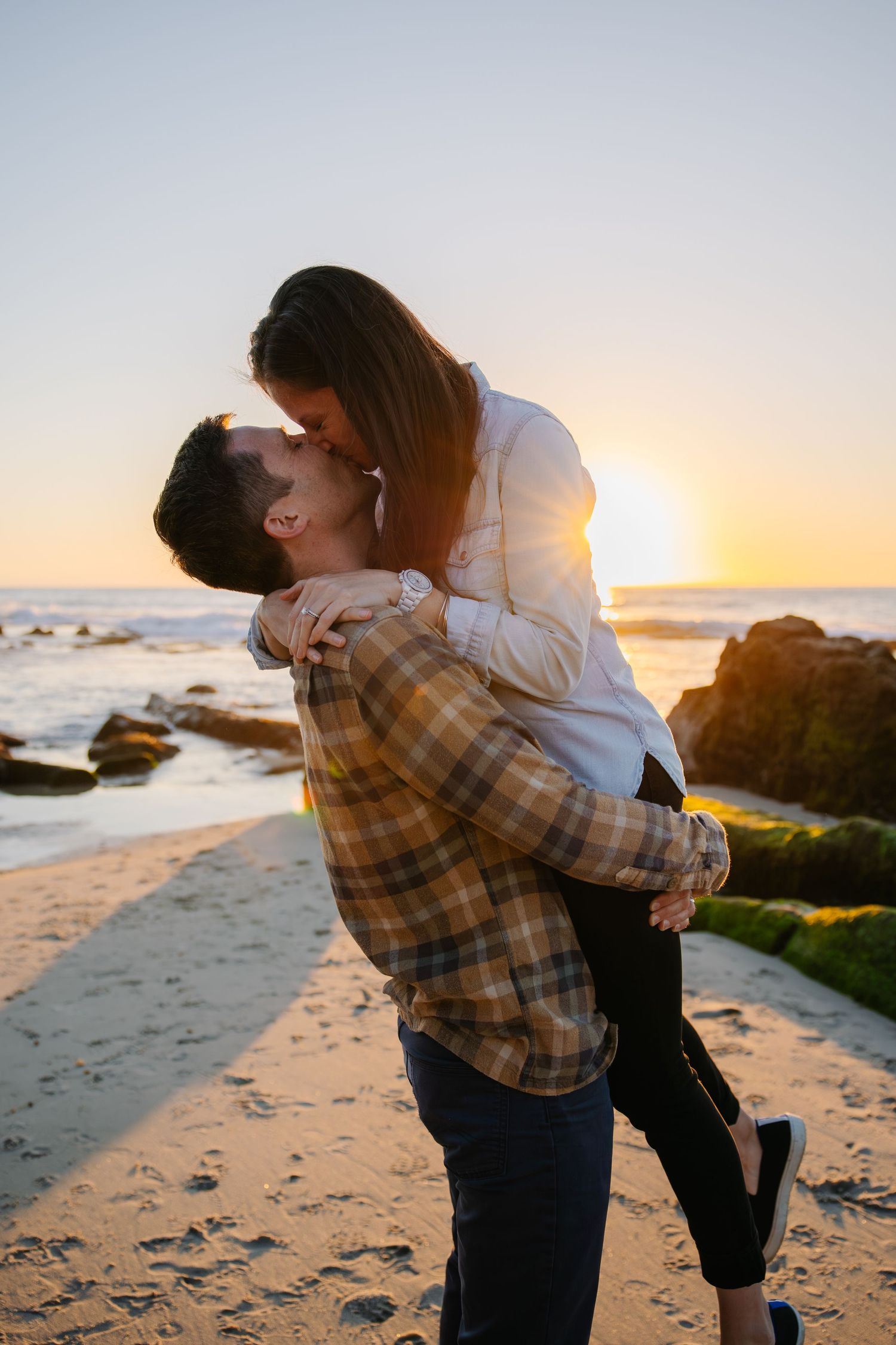 A romantic couple embraces on a beach at sunset with rocky shoreline in background.