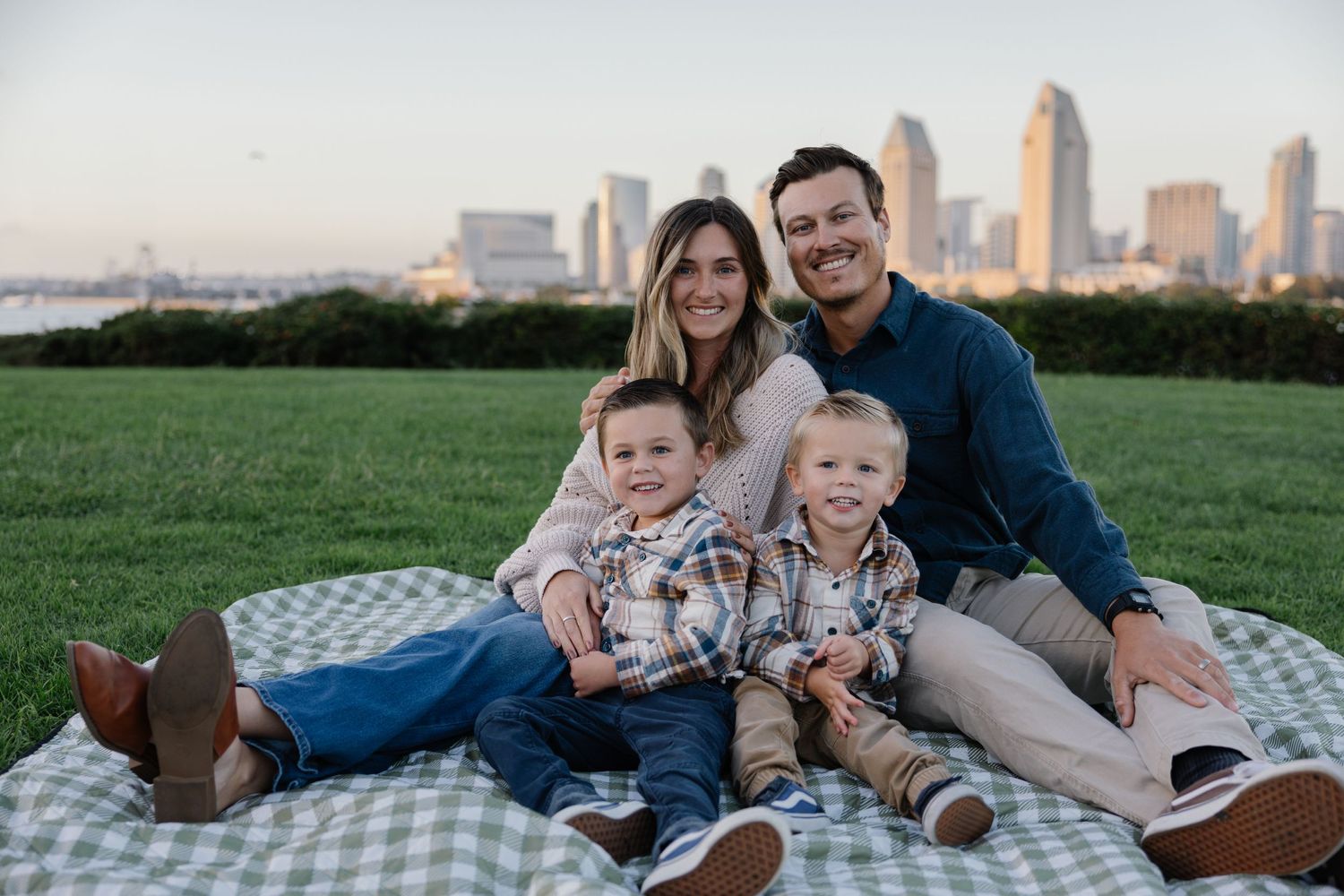 Family of four sitting on blanket for picnic with city skyline behind them.
