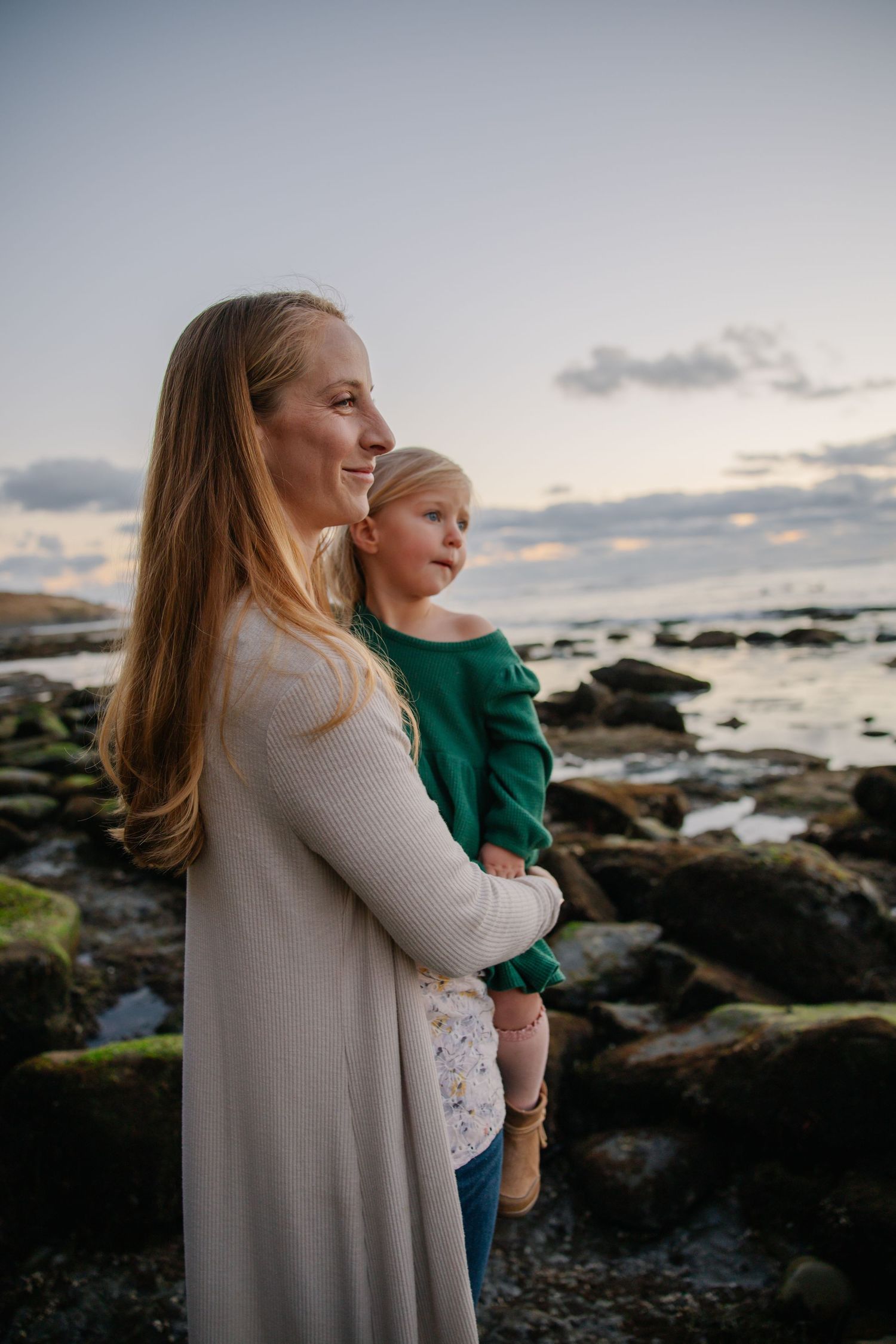 A parent and child stand together on rocky coastline during sunset.