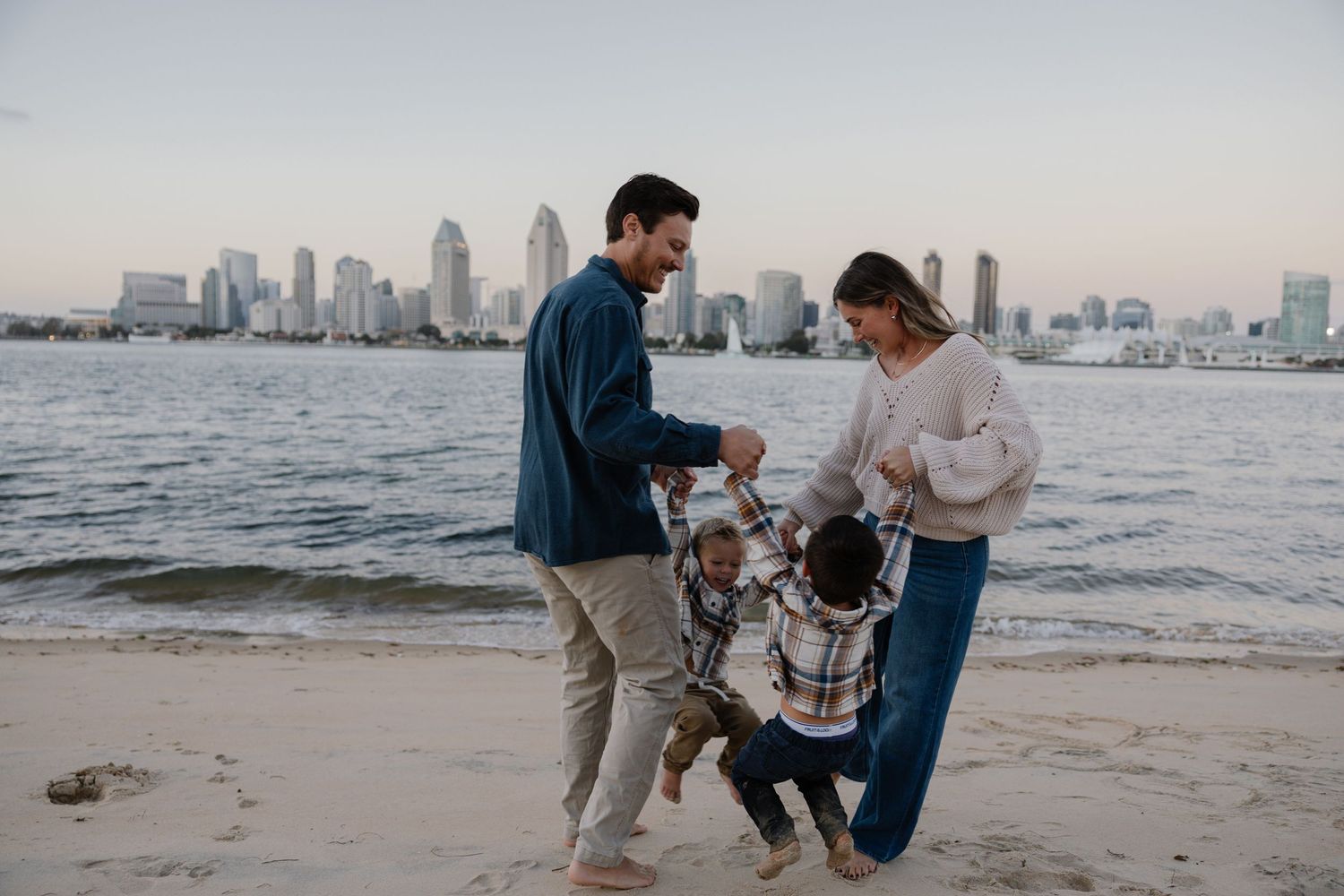 Family playing together on beach with city skyline in background at dusk.