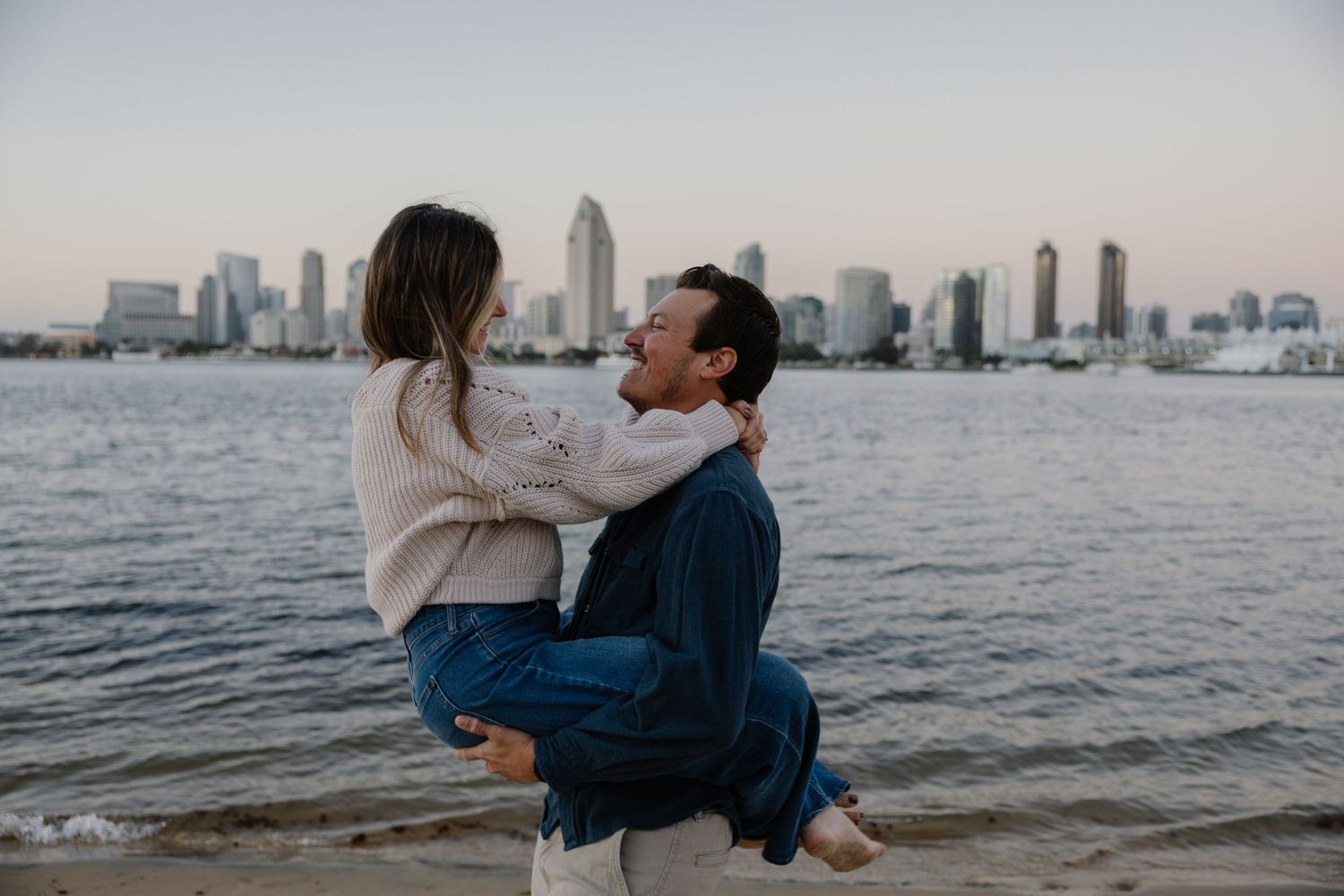 Couple embracing on beach with downtown skyline visible across water.