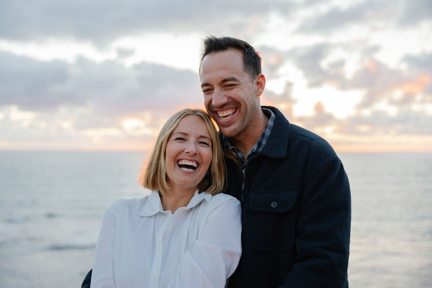 Happy couple laughing together at sunset by the ocean with cloudy sky in background.