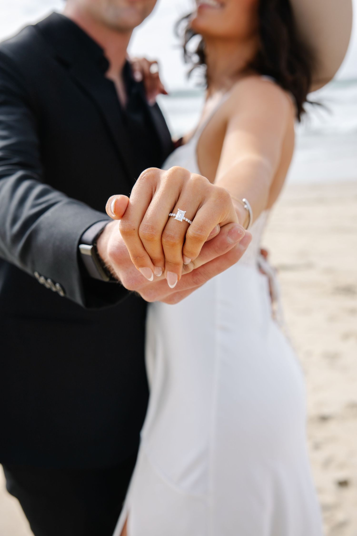 A close-up of an engagement ring as a couple holds hands on a beach.