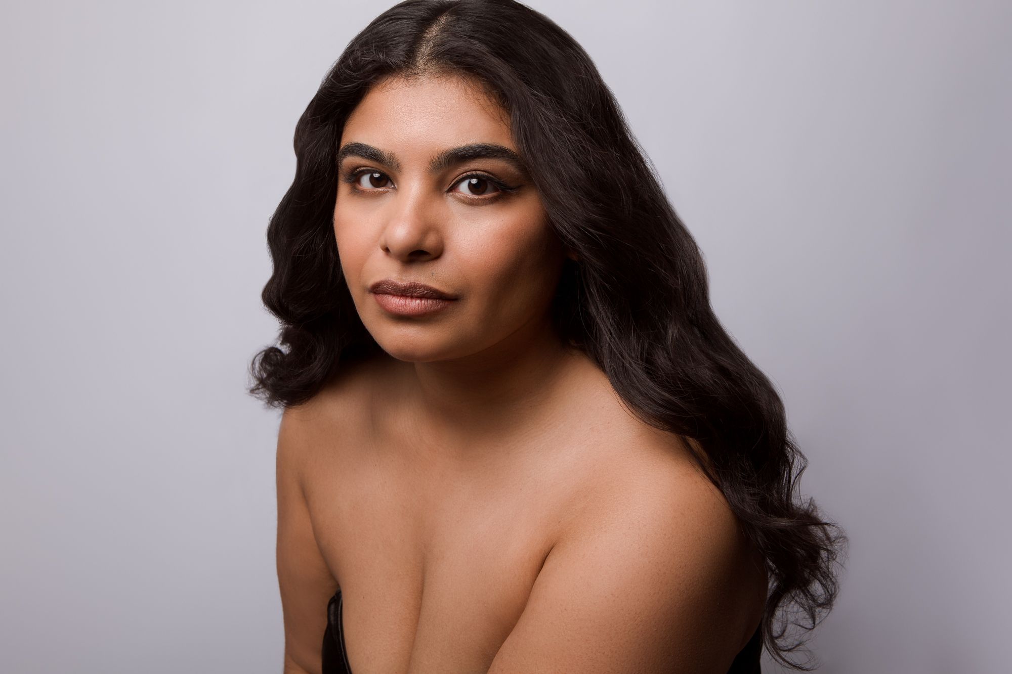 Studio portrait against gray background featuring long dark wavy hair and bare shoulders with natural makeup look.