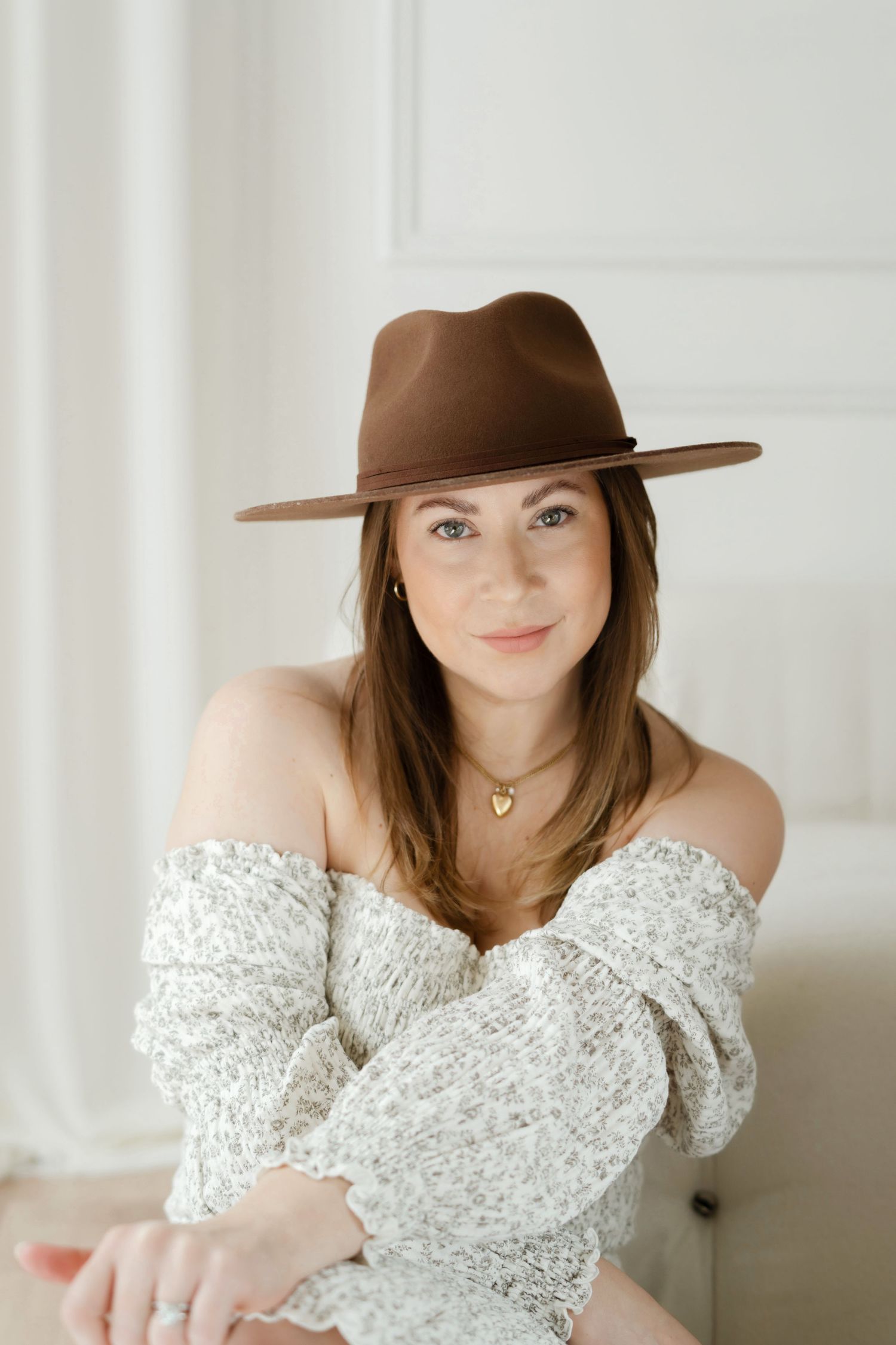 Elegant indoor portrait with brown hat and white off-shoulder dress against white wall.