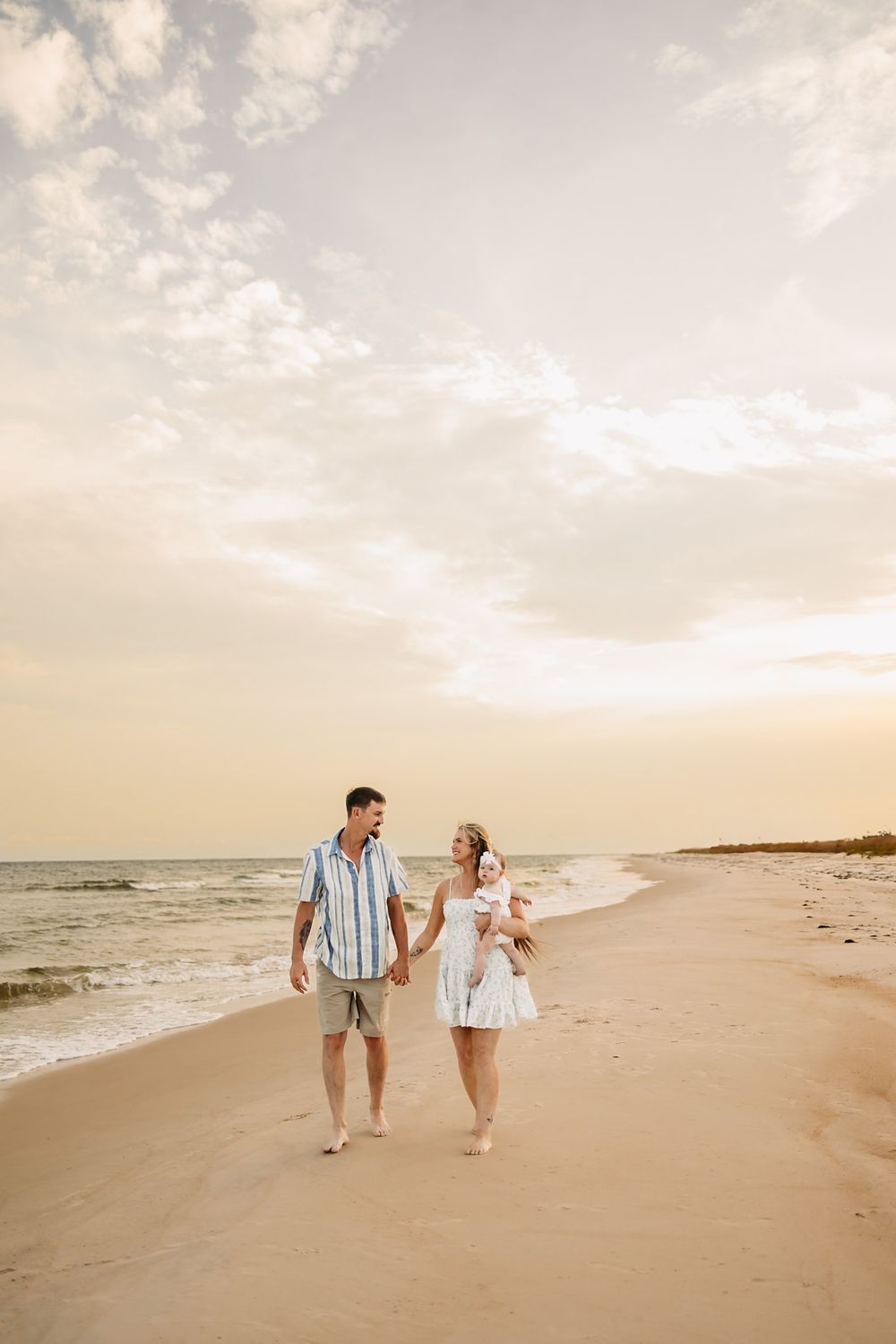 mom and dad holding hands walking down the beach with their baby on moms hip