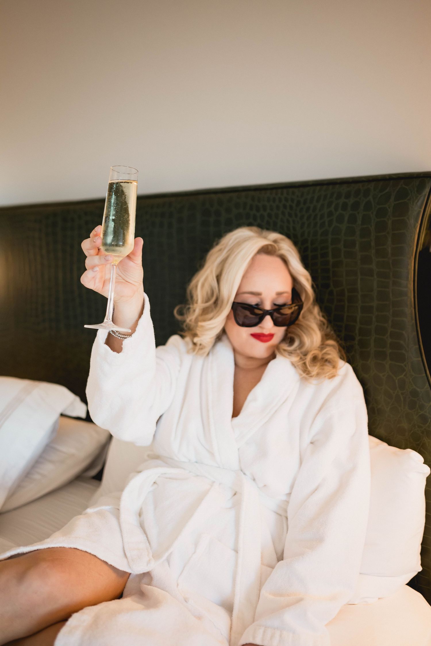 Person in white robe and sunglasses raising champagne glass while lounging in bed.