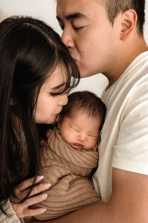 A sleeping newborn wrapped in a cozy brown knit blanket is tenderly embraced by their parents.