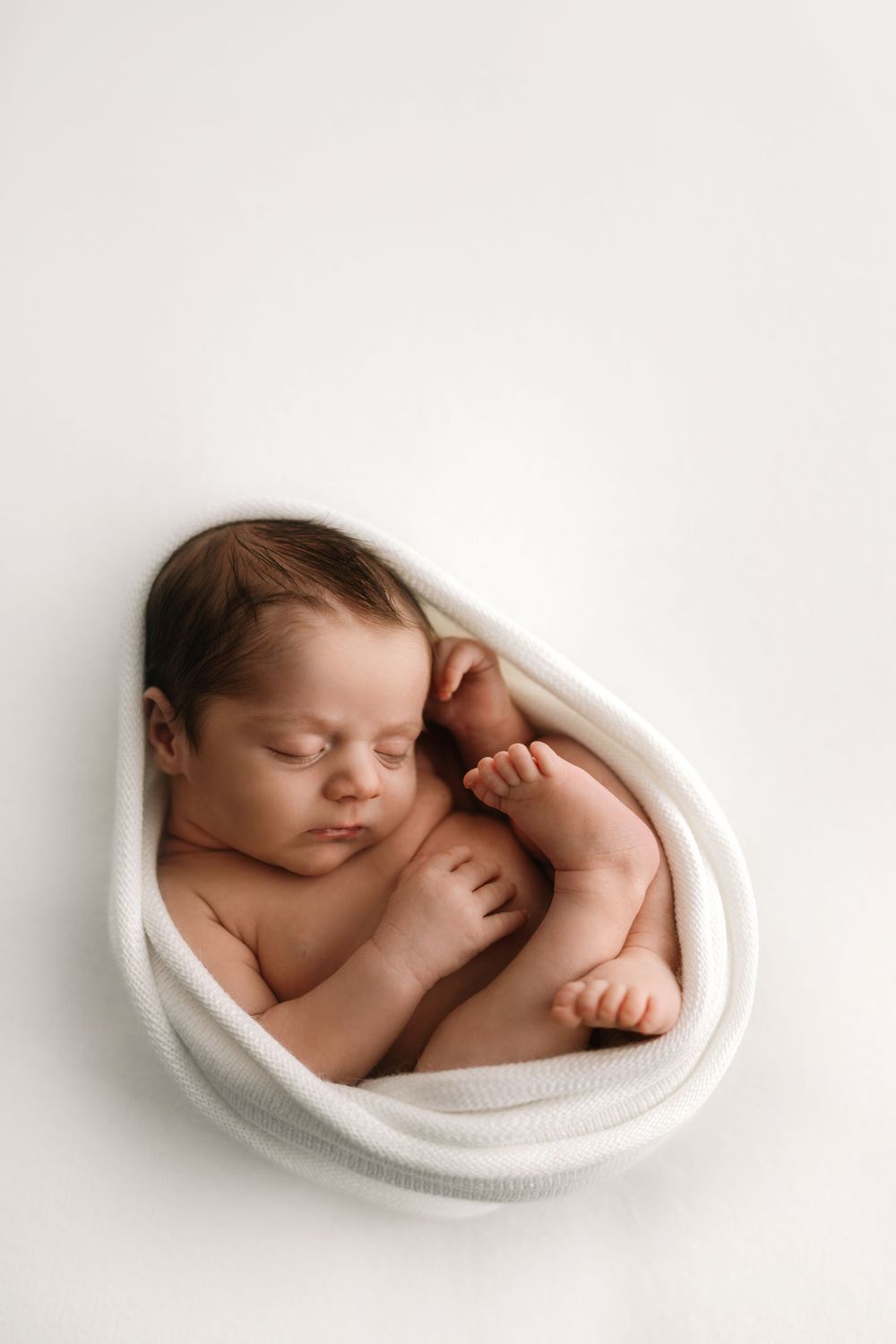 Peaceful sleeping newborn curled up in white wrap against minimalist background.