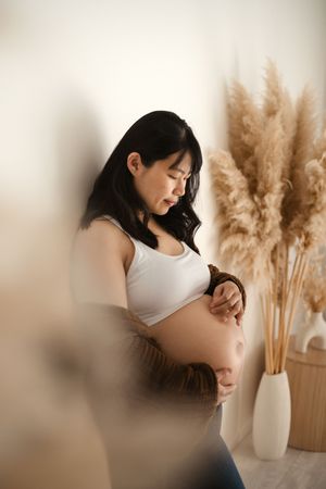 Artistic maternity photo series with white vase of pampas grass and soft neutral tones creating ethereal atmosphere.