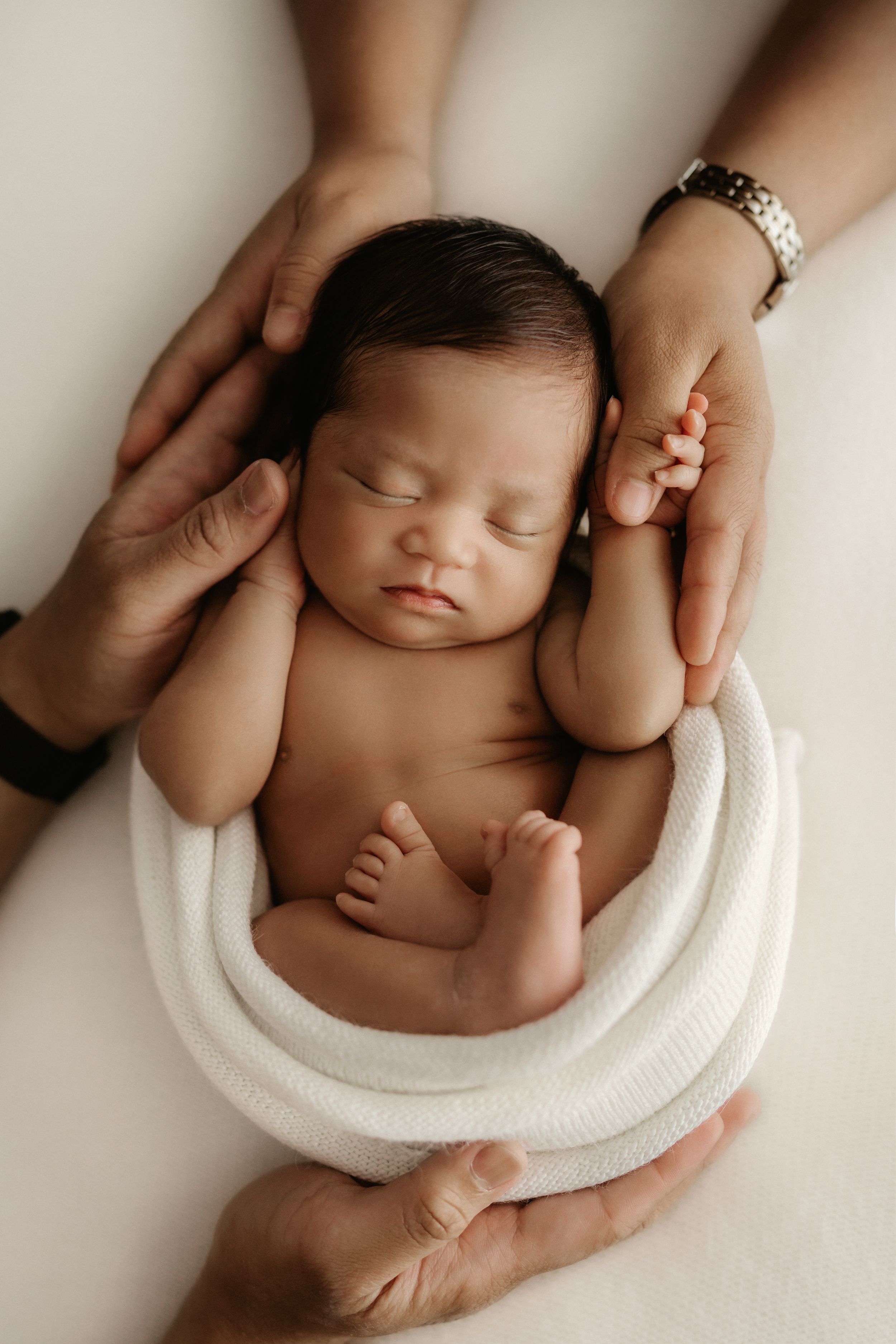 A sleeping newborn baby lies peacefully in a white blanket while gentle hands support them during a professional photo session.
