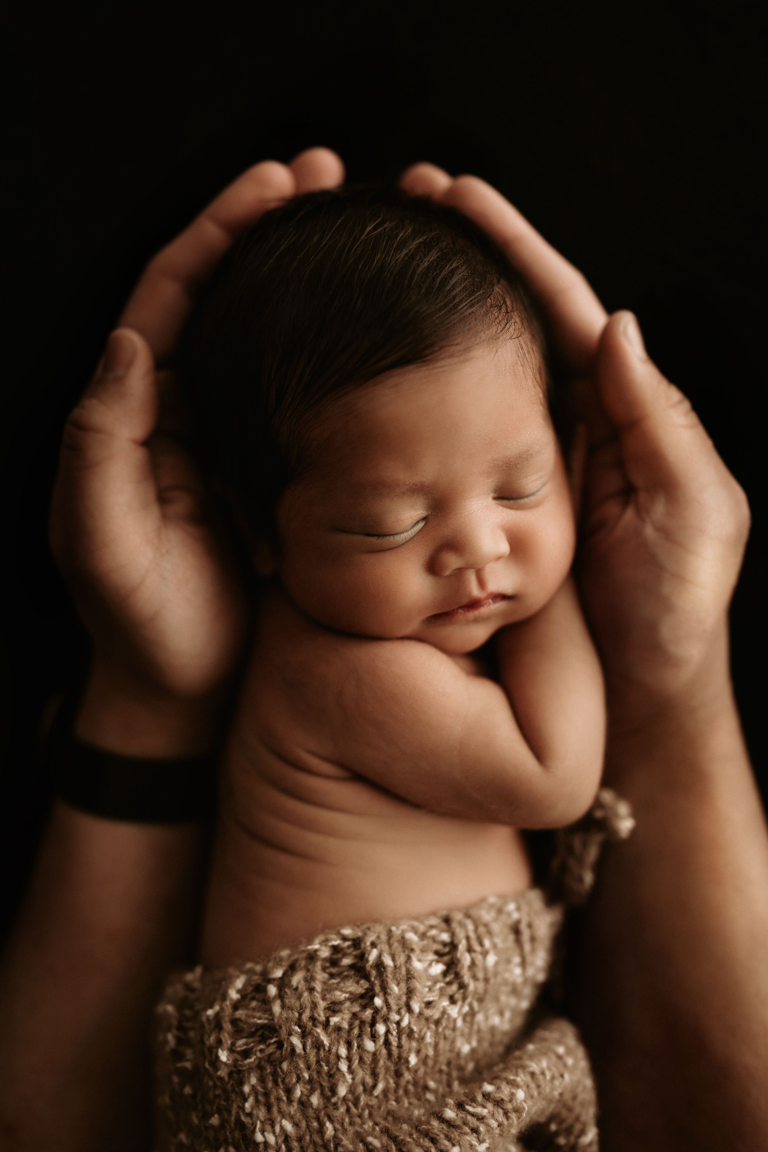 A sleeping newborn baby wrapped in gold fabric is gently cradled in adult hands against a dark background.
