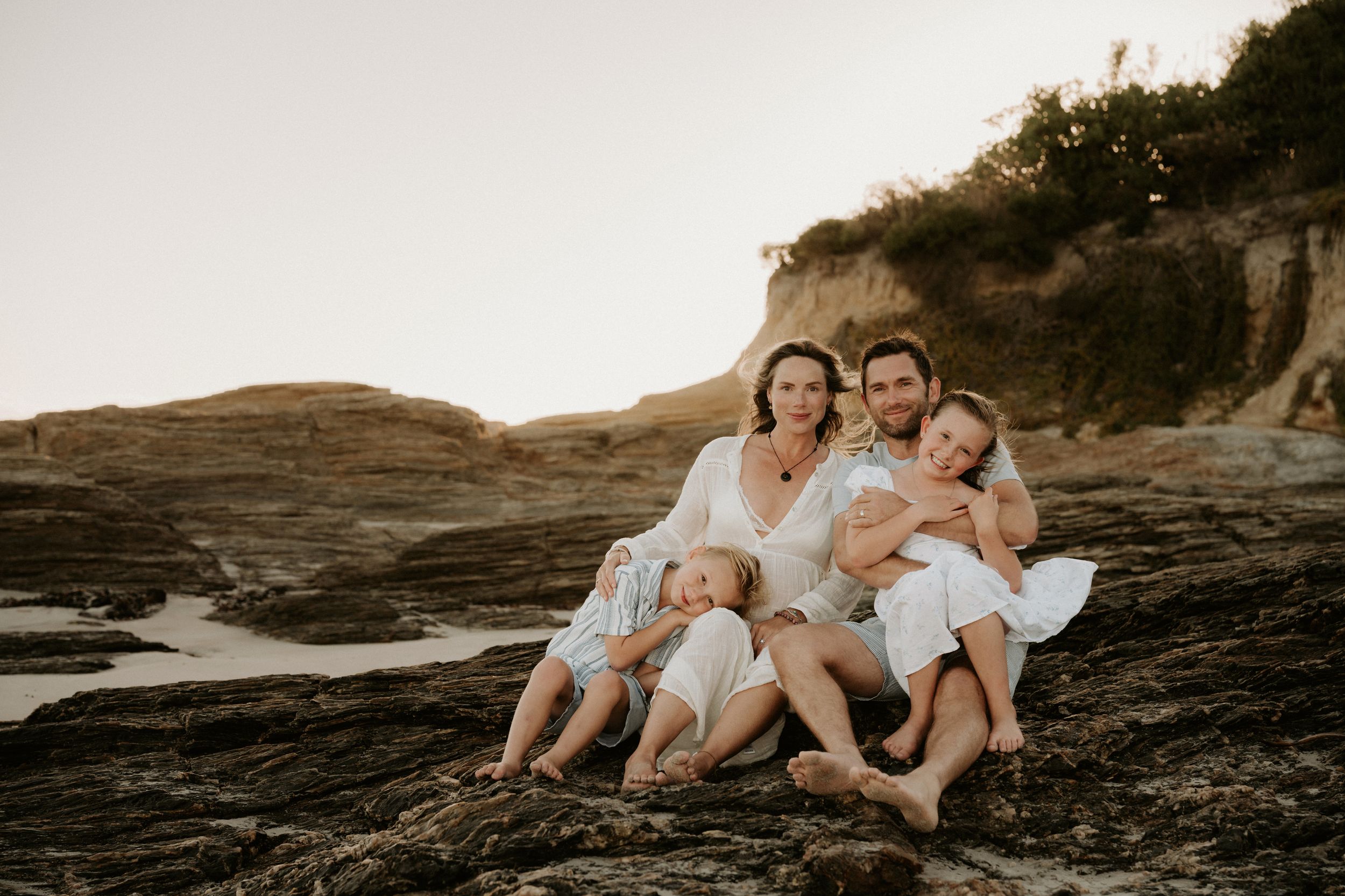 A family in white outfits sits together on rocky coastal cliffs during golden hour sunset.