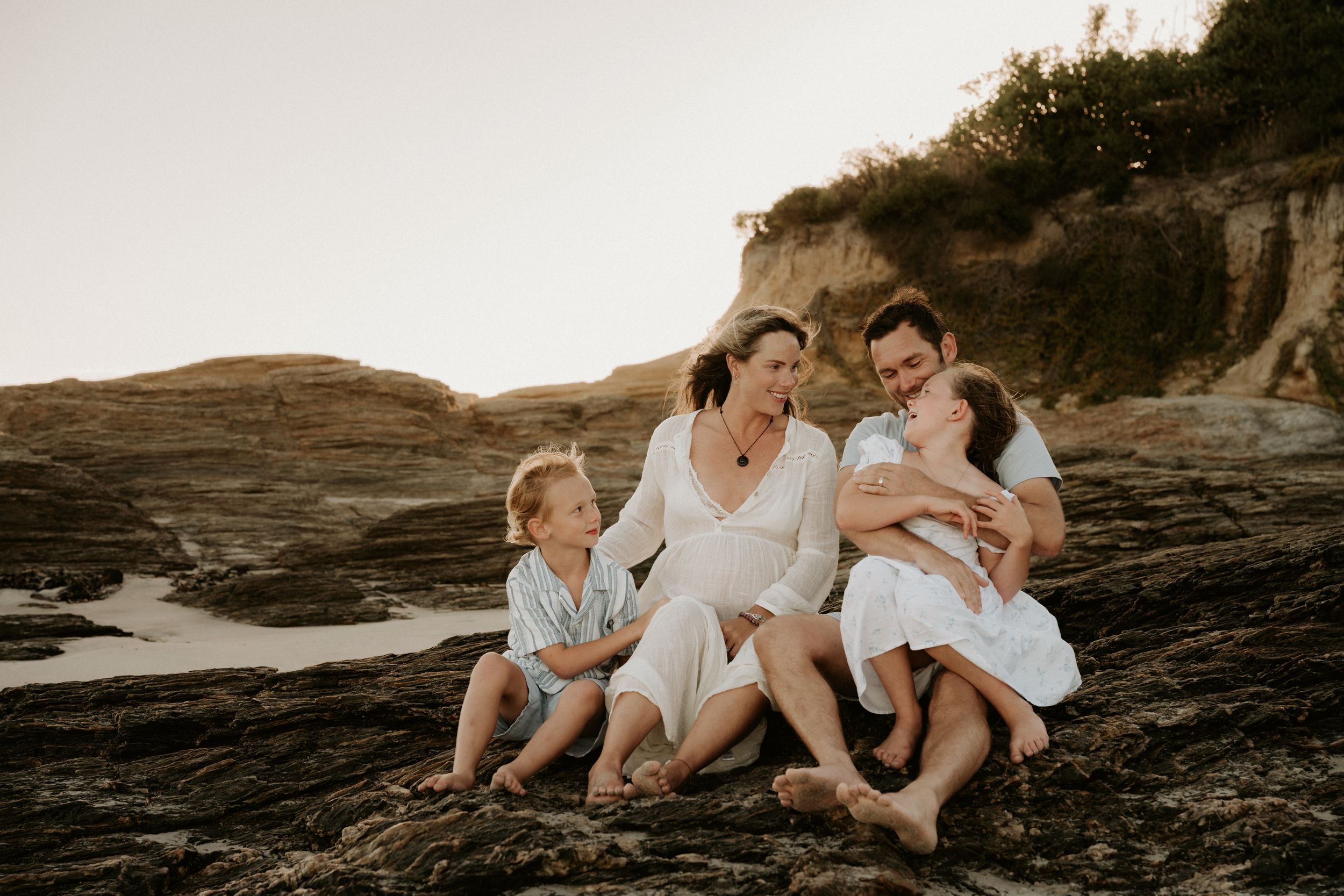A family sits together on rocky coastal terrain during golden hour sunset.