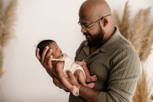 Bearded man wearing glasses and a sweater holds a newborn baby against a neutral backdrop with pampas grass.