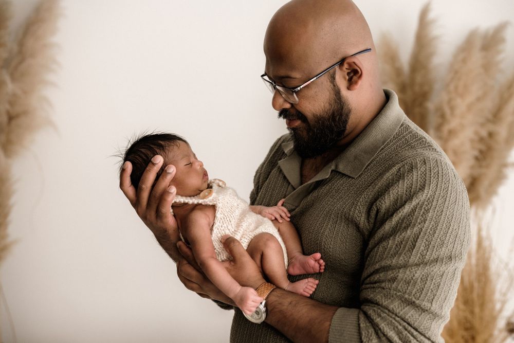 Bearded man wearing glasses and a sweater holds a newborn baby against a neutral backdrop with pampas grass.