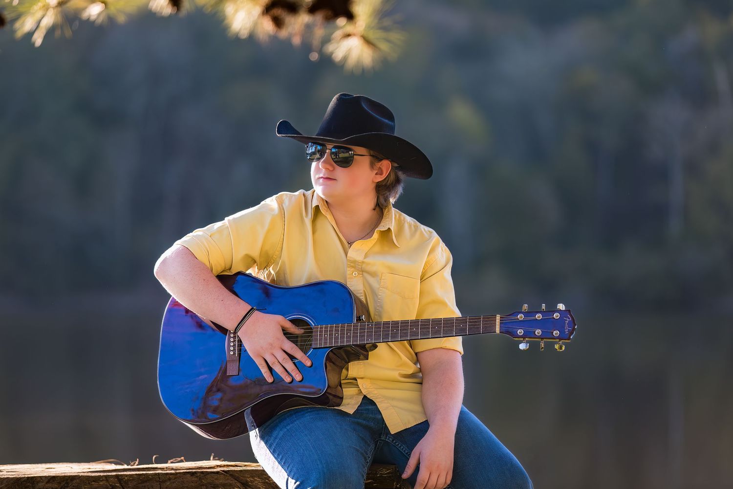 Country musician in yellow shirt and cowboy hat playing blue acoustic guitar while sitting by a lake.
